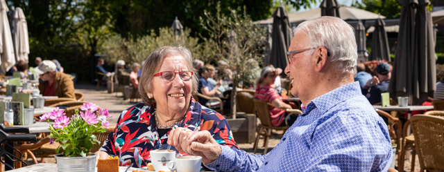 Ouder echtpaar zit op een zonnig terras met koffie en appeltaart.