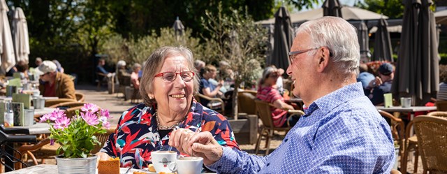 Ouder echtpaar zit op een zonnig terras met koffie en appeltaart.