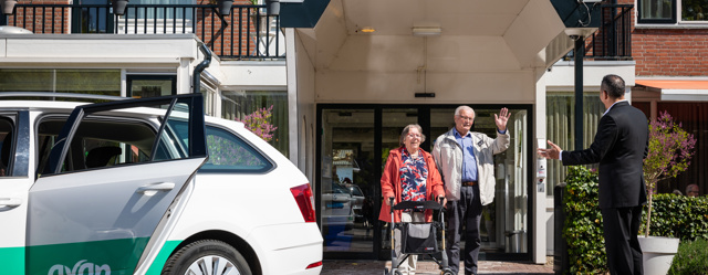 Echtpaar loopt naar buiten bij de woning en zwaait naar de Avan chauffeur hen op komt halen met een personenauto.