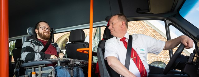 Een reiziger in een rolstoel zit in de taxibus, de chauffeur kijkt om naar de reiziger.